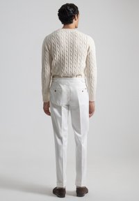 Suéter de punto trenzado crema, pantalones blancos ajustados con bolsillos traseros y zapatos de ante marrón, fotografiados desde atrás contra un fondo neutral.