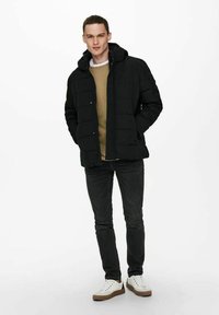 Puffer jacket in black with a hood, featuring a quilted design. Worn over a beige sweater, paired with dark jeans and white sneakers.