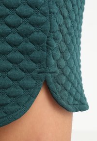 Tejido acolchado en color azul verdoso con patrón de diamantes y una pequeña hendidura que revela la piel debajo, sugiriendo un detalle en el dobladillo de la prenda.