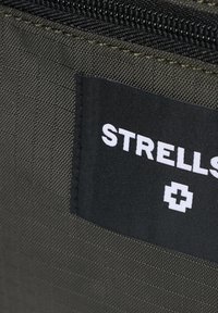 Un tessuto verde con una superficie texture presenta un'etichetta nera con testo bianco che recita "STRELLS" e un simbolo di croce bianca.