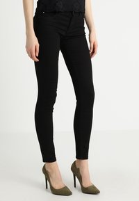 Jeans skinnies negros con una textura suave, cintura alta y cinco bolsillos, combinados con zapatos de tacón altos en color verde oliva.