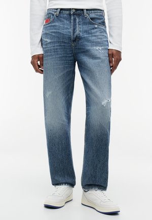 Homme portant un jean bleu délavé droit avec une légère usure, des baskets blanches et une chemise blanche à manches longues, debout sur un fond uni.