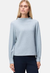 Top azzurro chiaro con texture, colletto alto e maniche lunghe, caratterizzato da un motivo a diamante sottile. Indossato con pantaloni scuri.
