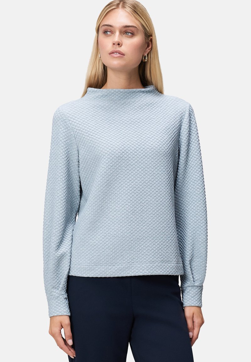 Top azzurro chiaro con texture, colletto alto e maniche lunghe, caratterizzato da un motivo a diamante sottile. Indossato con pantaloni scuri.