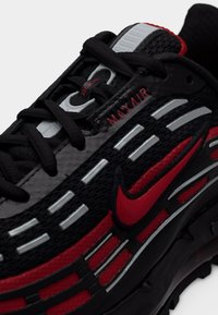 Chaussure de sport noire et rouge avec une tige en mesh, des accents argentés, un amorti visible à l'air et des lacets noirs. Présente un design courbé et un détail de logo.