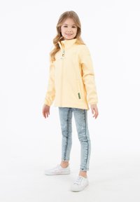 Geltonas vandeniui atsparus švarkas su aukštu apykakle, užtrauktuku priekyje ir dviem šoninėmis kišenėmis. Suderintas su šviesiai mėlynomis džinsomis ir raštuotais sportbačiais.