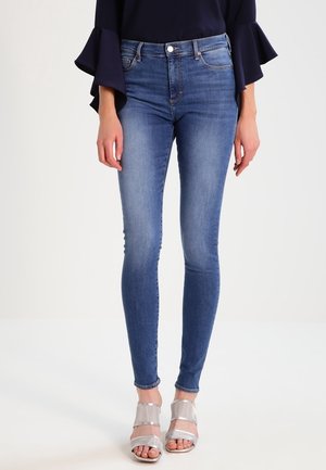 Femme portant un jean skinny bleu, un haut bleu marine à manches évasées et des sandales argentées à talons ouverts sur les doigts, debout devant un fond blanc.