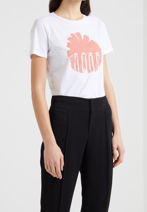 Kobieta ubrana w białą koszulkę z różowym nadrukiem i napisem „ROSS” w połączeniu z czarnymi spodniami wysokiego stanu o kroju na miarę na jednolitym tle.