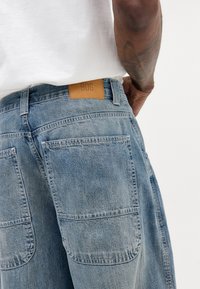 Hellblaue Denim-Jeans mit einem Lederschild von BDG am Bund, ausgestattet mit zwei Gesäßtaschen und einer leicht verblassten Textur.