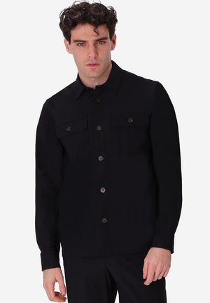 Uomo con capelli ricci e scuri che indossa una camicia nera con bottoni e tasche con patta, in piedi davanti a uno sfondo bianco.