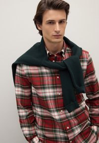 Rotes Karohemd mit weißen und grünen Akzenten, kombiniert mit einem dunkelgrünen Strickpullover, der lässig über die Schultern gelegt ist. Schlichter, legerer Stil.