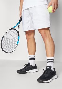Tennis-Outfit mit weißen Textur-Shorts, schwarzen Turnschuhen, schwarz-weißen gestreiften Socken und einem blau-schwarzen Tennisschläger, der einen gelben Ball hält.