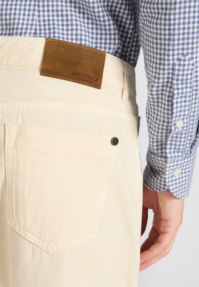 Pantalones de algodón beige con un bolsillo trasero doblado, parche de ante marrón etiquetado "INCOTEX" y una camisa de cuadros azules con puños de botón.
