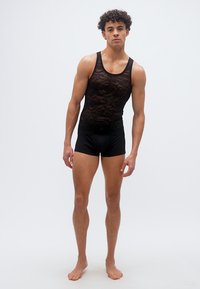 Νεαρός άνδρας σε όρθια θέση, ξυπόλητος, φοράει μαύρο δαντελένιο αμάνικο τοπ και μαύρο εφαρμοστό σορτς, σε απλό ανοιχτό φόντο.