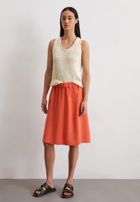 White knit tank top and an orange knee-length skirt. Black sandals with gold buckle accents. Smooth textures and relaxed fit.