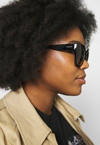 Óculos de sol oversized pretos com um acabamento brilhante, apresentando o nome da marca em relevo "vogue" na lateral, usados por uma pessoa com cabelo natural.