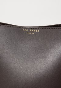 Sac en cuir texturé marron avec le logo embossé "Ted Baker London" en doré. Fini lisse et éclat subtil ; design minimaliste sans matériel visible.