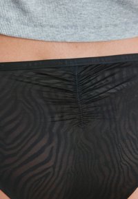 Czarne bikini dolne z przezroczystym, teksturowanym wzorem w zebrę, z marszczeniem z tyłu. Widoczny elastyczny pas.