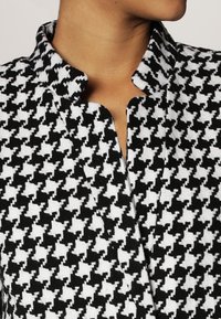 Svart och vit hounds-tooth-mönstrad tyg med krage och öppen halsringning, som visar en strukturerad väv och växlande former.