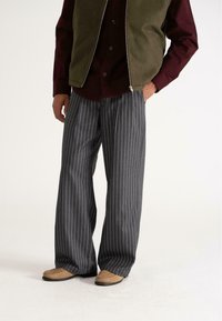 Uomo che indossa pantaloni larghi a righe verticali grigio scuro, mocassini in suede marroni, camicia bordeaux e gilet verde oliva con zip, in piedi con le mani in tasca.