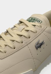 Baskets en cuir beige avec lacets plats, logo Lacoste en argent et détails perforés le long des côtés. Surface légèrement texturée.