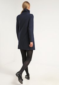 Cappotto di lana navy con colletto alto, design sartoriale, maniche lunghe e lunghezza fino a metà coscia. Indossato con collant neri e scarpe.