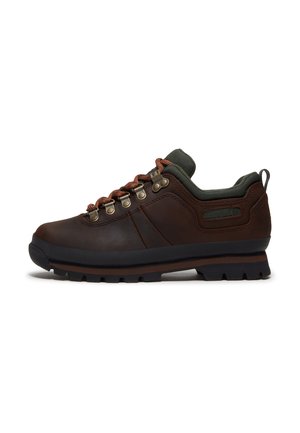 Zapato de senderismo bajo marrón con cordones naranjas, ojales de metal, cuello acolchado y suela de goma negra sobre fondo blanco.