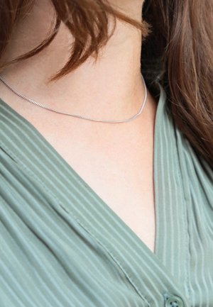 Zilveren ketting met een fijne, flexibele chain rustend op een blote nek, gecombineerd met een groen gestreepte blouse. Soepele textuur, minimalistisch ontwerp.