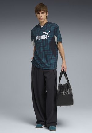 Młody mężczyzna w turkusowej sportowej koszulce Puma, luźnych czarnych spodniach, turkusowych butach, trzymający czarną skórzaną torebkę na tle szarego tła.