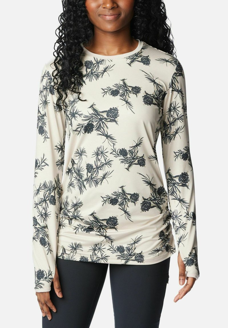 Frau trägt ein beiges Langarmshirt mit schwarzem botanischem Muster und Daumenlöchern, kombiniert mit schwarzen Leggings vor einem schlichten weißen Hintergrund.