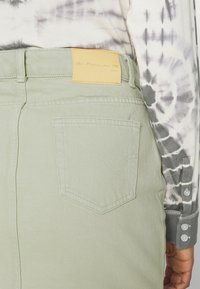 Gonna di jeans verde chiaro sul retro con una tasca cucita e un'etichetta del marchio beige, abbinata a una maglietta a maniche lunghe bianca e grigia con effetto tie-dye, risvoltata ai polsi.