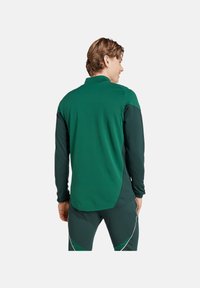 Maglietta sportiva verde a maniche lunghe caratterizzata da una vestibilità sagomata, tessuto testurizzato e pannelli a contrasto sulle spalle. Presenta un orlo curvo e un design elegante.