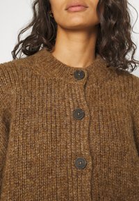 Brun stickad cardigan med rund halsringning, prydd med tre mörka knappar och ett strukturerat, tjockt tyg.