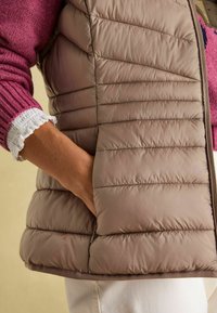 Beige Steppweste mit vertikalen Nähten, ausgestattet mit einer seitlichen Tasche und einem gerippten Bündchen aus einem Kontrast-Pink bei einem Strickpullover.