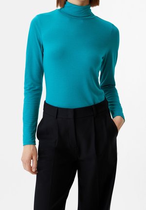 Personne portant un haut côtelé à col roulé turquoise à manches longues et un pantalon noir taille haute, une main dans la poche, sur un fond uni.