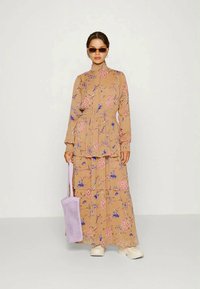 Vestido maxi floral em bege com camadas volumosas, mangas longas, um decote alto e padrões de flores roxas e cor-de-rosa. Acompanhado de uma bolsa lavanda.