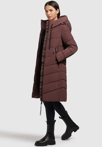 Dlouhý burgundský puffer kabát s kapucí, s zipem a knoflíkovým zapínáním, ve tvaru šípů prošívaný povrch a dvě boční kapsy. Viditelné černé boty.