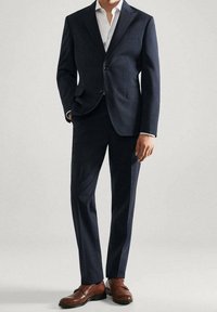 Costume bleu marine avec une texture, veste à un bouton et pantalon droit. Associé à des chaussures en cuir marron et une chemise blanche.