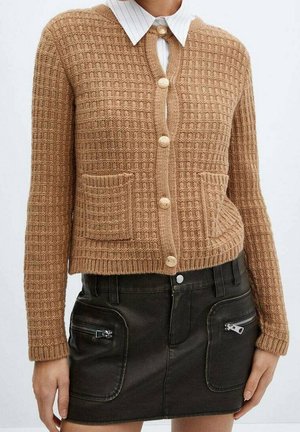 Cardigan en tricot couleur fauve avec boutons sur le devant et deux poches, porté sur une chemise blanche à col, associé à une mini-jupe noire avec poches zippées.