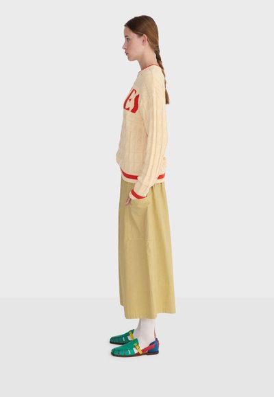 Jeune femme en pull beige avec des lettres rouges, longue jupe beige, chaussettes blanches et sandales colorées vertes, jaunes et bleues, debout de profil.