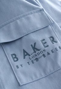 Svetlomodrá tkanina s hladkou textúrou s vystúženou vreckom. Vrecko nesie embosovaný nápis: "BAKER by Ted Baker."