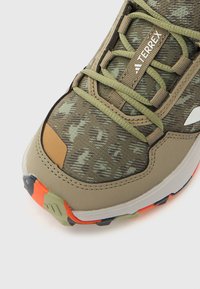Πράσινο και μπεζ αθλητικό παπούτσι με υφή στην επάνω επιφάνεια, κλείσιμο με κορδόνια, πορτοκαλί και μαύρες λεπτομέρειες και διακοσμητικό σχέδιο στο ύφασμα.