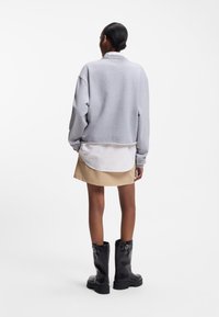 Γυναίκα που στέκεται με την πλάτη προς τα επάνω, φορώντας γκρι φούτερ, λευκό πουκάμισο, μπεζ φούστα και μαύρες μπότες μέχρι τη μέση της γάμπας, με φόντο λευκό.