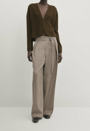 Osoba w brązowym rozpinanym kardiganie i beżowych, wysokim stanem, szerokich spodniach z pasem do kompletu, stojąca na jednolitym tle.