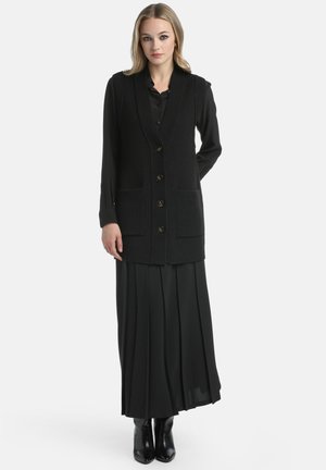 Woman wearing a black long-sleeve shirt, black buttoned knit vest with pockets, black pleated maxi skirt, and black ankle boots, standing.