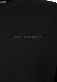 Suéter de punto negro con un cuello acanalado. Presenta un logo negro texturizado "KARL LAGERFELD" centrado en el frente. Diseño simple, sin patrones.