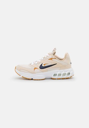 Baskets Nike beige et blanches avec un swoosh noir, des accents orange, des lacets blancs et un amorti visible dans la semelle, sur un fond neutre.