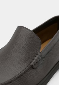 Tmavosivá textúrovaná kožená slip-on obuv so šitými detailmi a hladkým zaobleným špičkou, zobrazená zblízka na bielom pozadí.