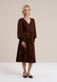 Modellen står i en bourgogne-färgad blommig klänning med långa puffärmar, åtdragen vid midjan, tillsammans med svarta skor, mot en neutralt bakgrund.