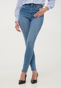 Dżinsy skinny w kolorze niebieskim z wysokim stanem, wykonane z denimu, charakteryzujące się dopasowanym krojem, wyraźnymi szwami i surowym dołem. Połączone z czarnymi szpilkami w szpic.
